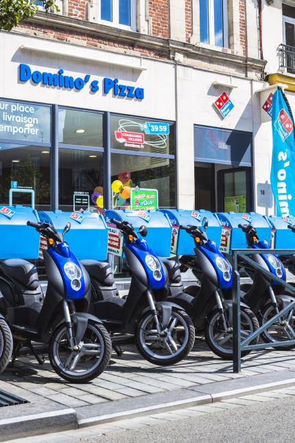 Façade d'un Domino's Pizza - enseigne reconnaissable aux couleurs bleu ciel et rouge -, avec juste devant les scooters utilisés pour la livraison à domicile.